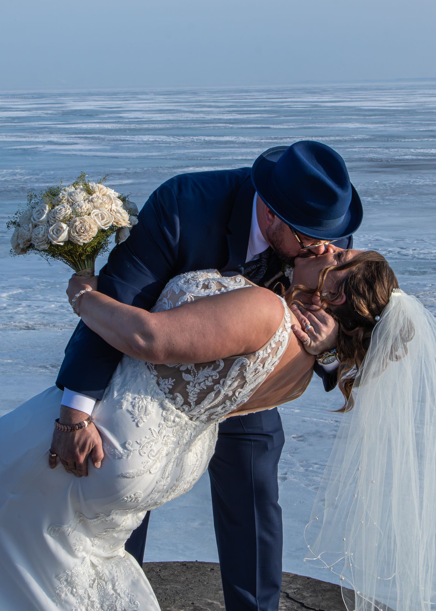 A romantic wedding couple shares a dip kiss on a beach with ocean waves in the background.