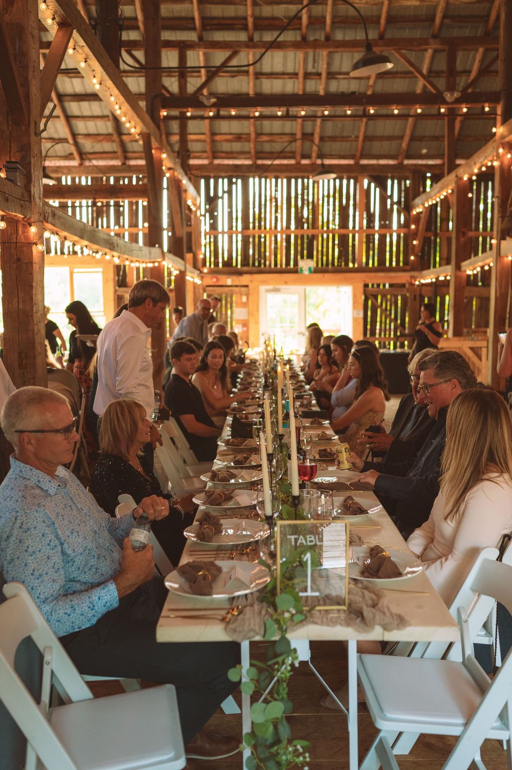 Guests seated at long rustic dining tables inside a barn with string lights and natural light filtering through wooden slats.