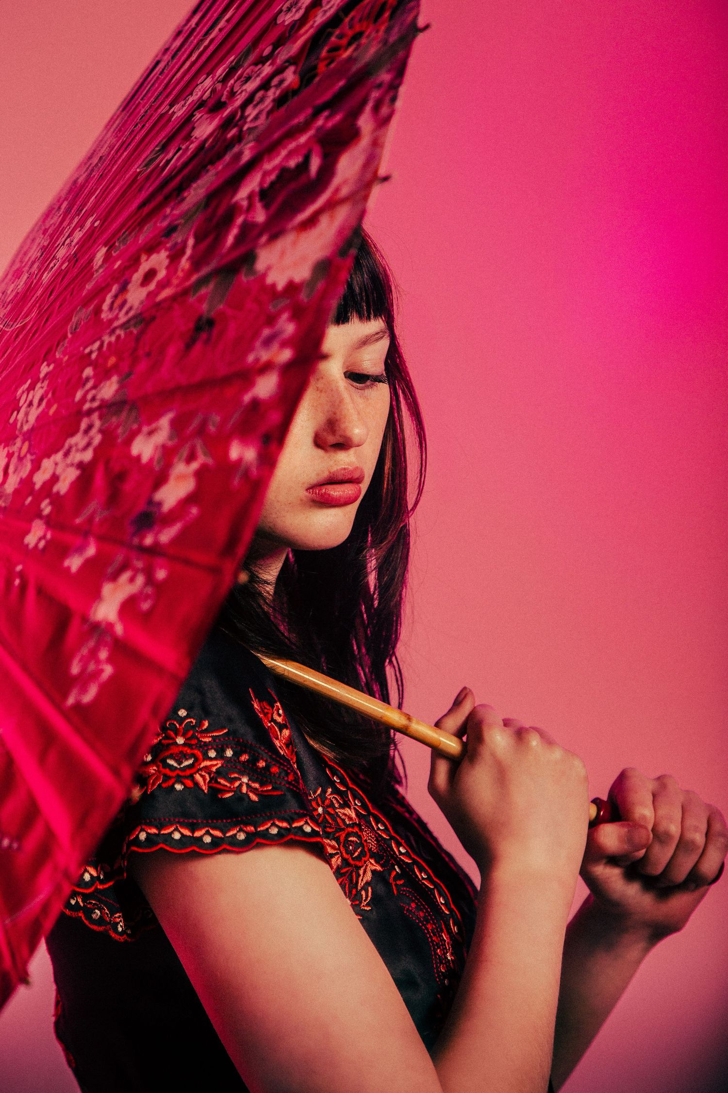 A person in traditional Asian attire holds a red decorative umbrella against a pink background in dramatic lighting.