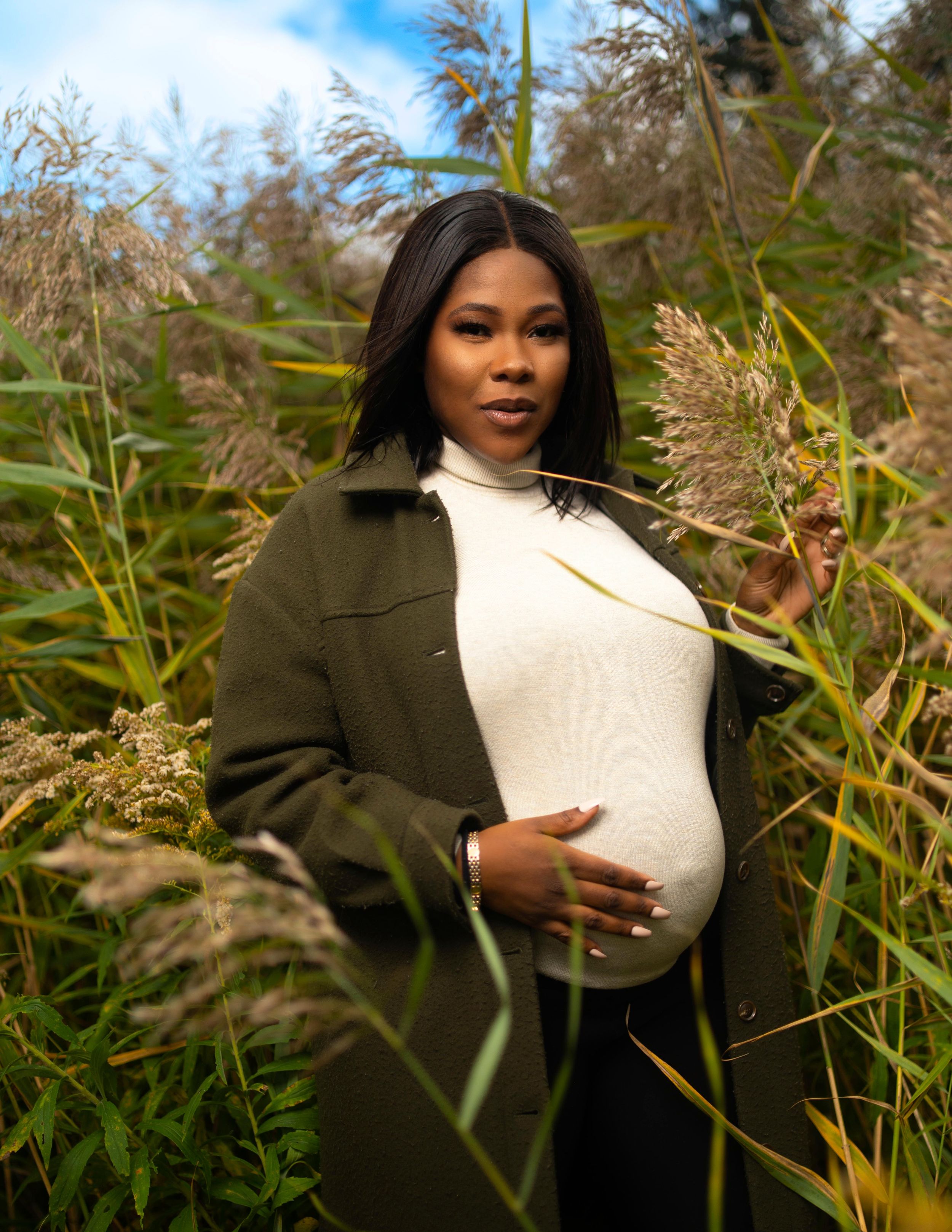 A pregnant woman in a white sweater and olive coat cradles her bump in a field of tall grass.