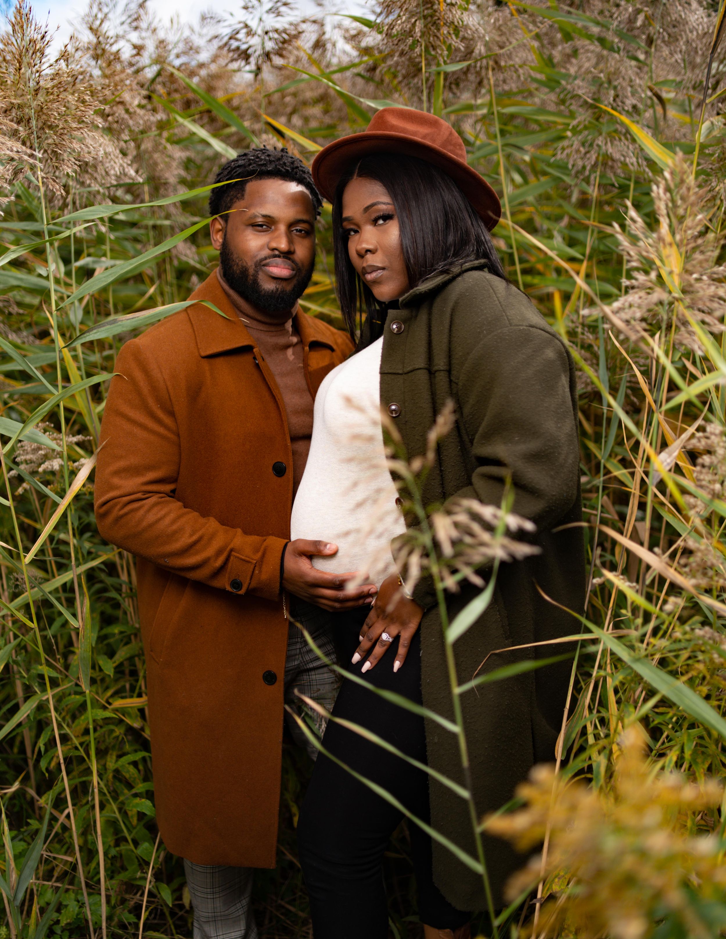 A couple poses together in a field of tall grass during an outdoor fall maternity photoshoot.