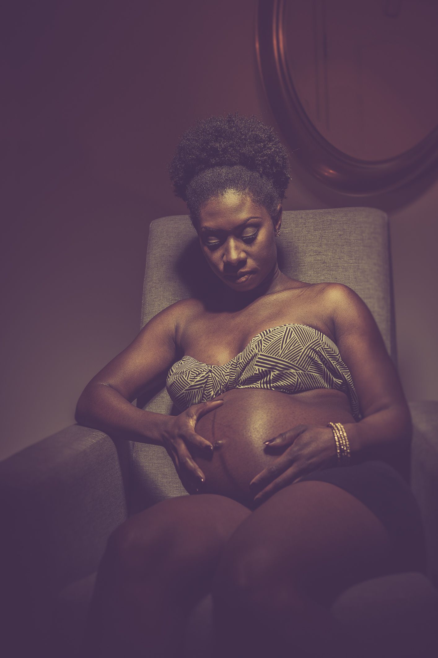 A pregnant woman in a gold bandeau top cradles her baby bump while seated in a chair.
