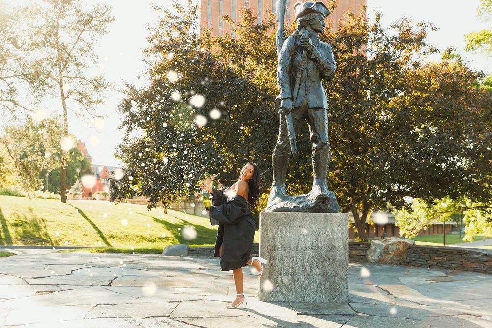 Graduate posing near bronze statue on university campus during sunset.