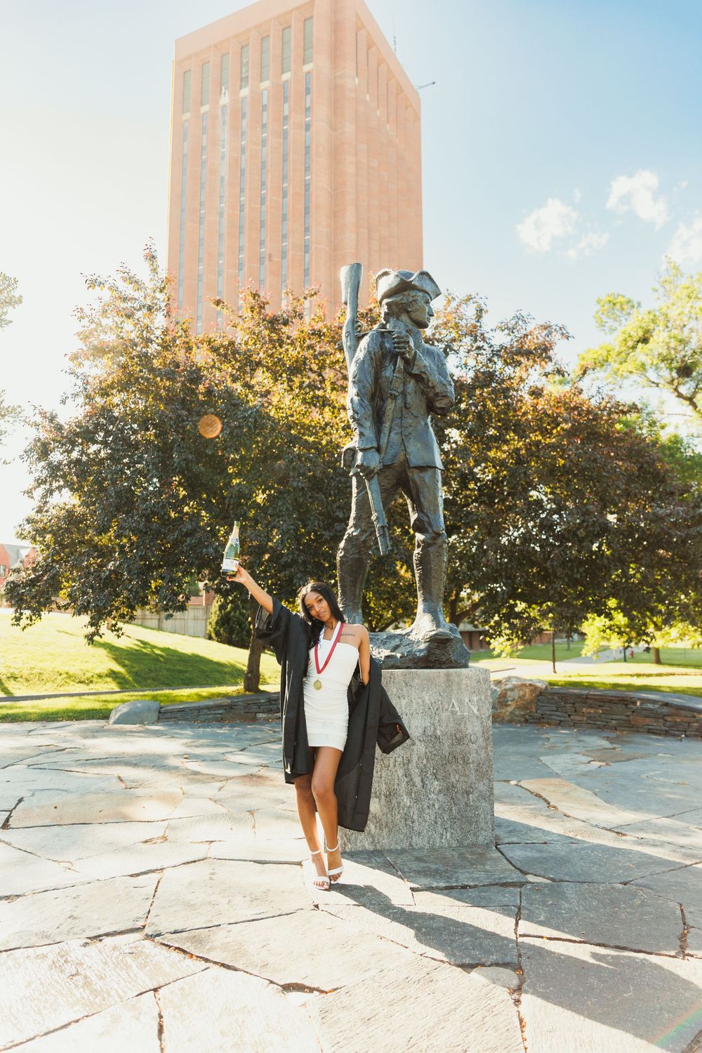 Graduate celebrating by campus statue and brick building on graduation day.