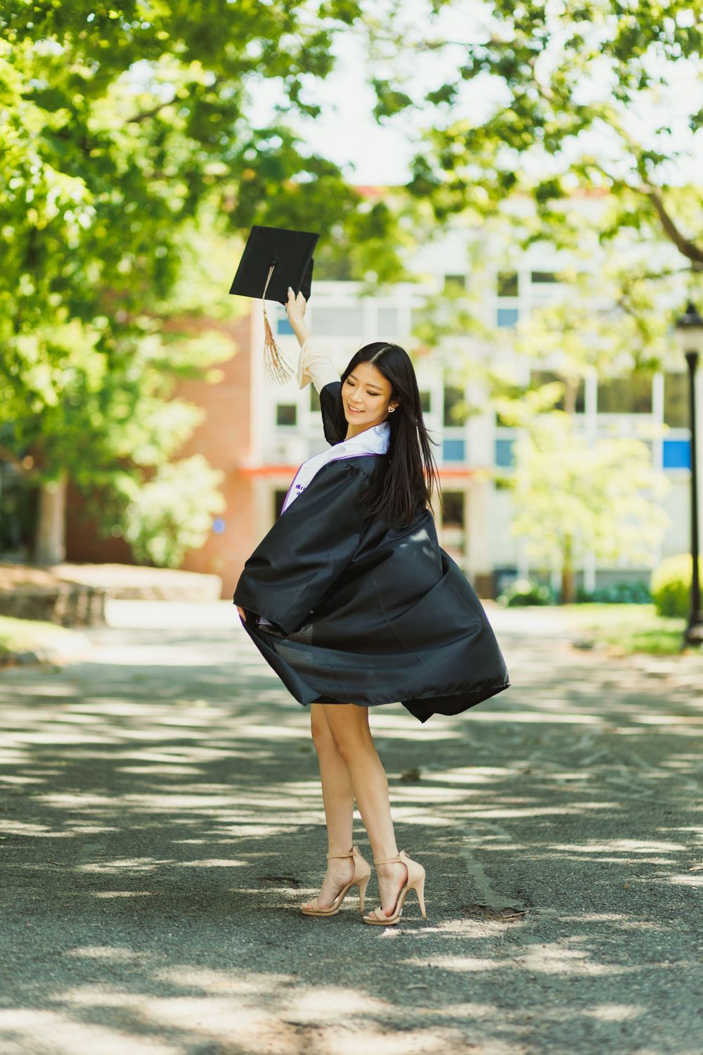 Graduate in black cap and gown twirling on campus pathway.