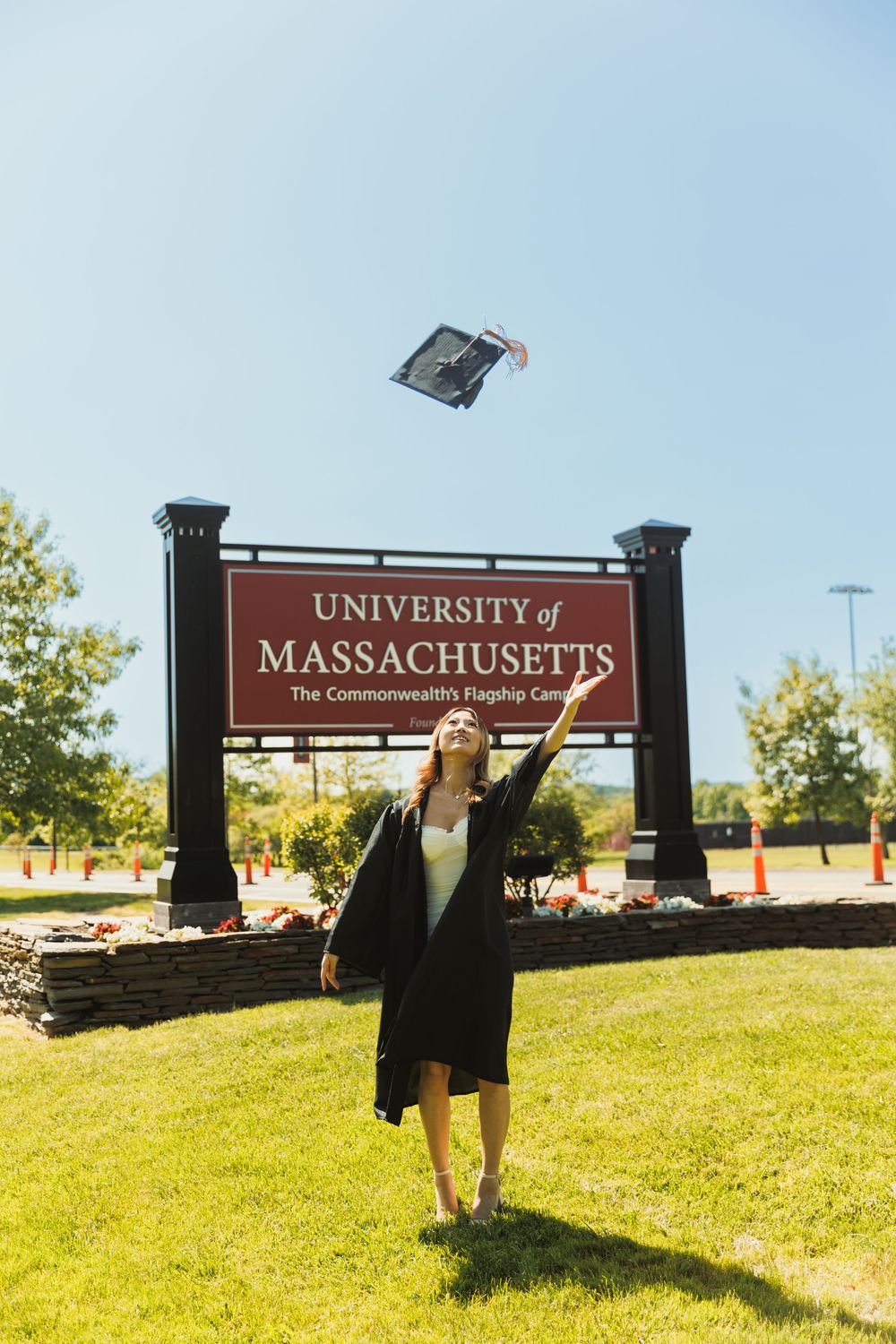 Graduate tossing cap in front of University of Massachusetts campus sign.