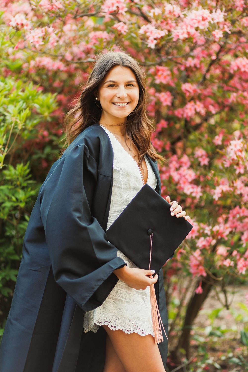 Graduate holding diploma poses in front of flowering pink tree in traditional black graduation gown.
