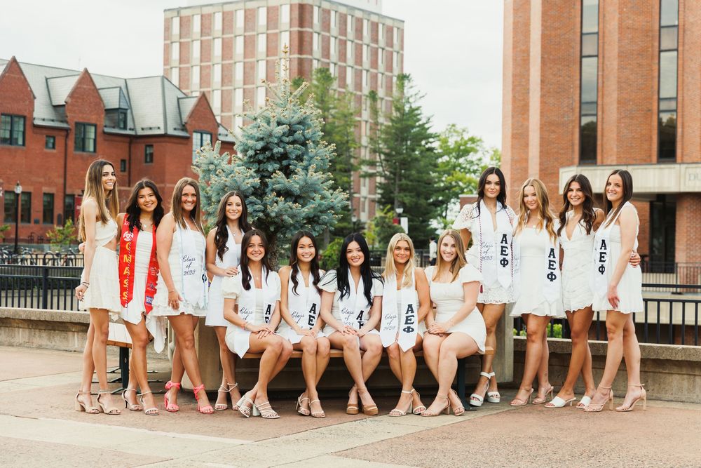 Sorority sisters in white dresses pose together for graduation photo on campus.