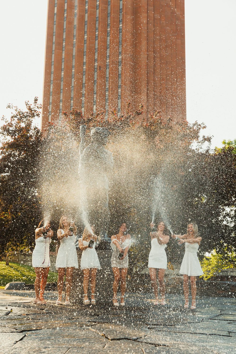 Graduates celebrating with water splashes in front of a brick university building.