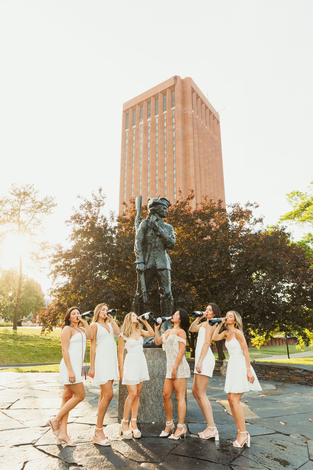 Graduates posing together in front of a campus statue and brick building at sunset.