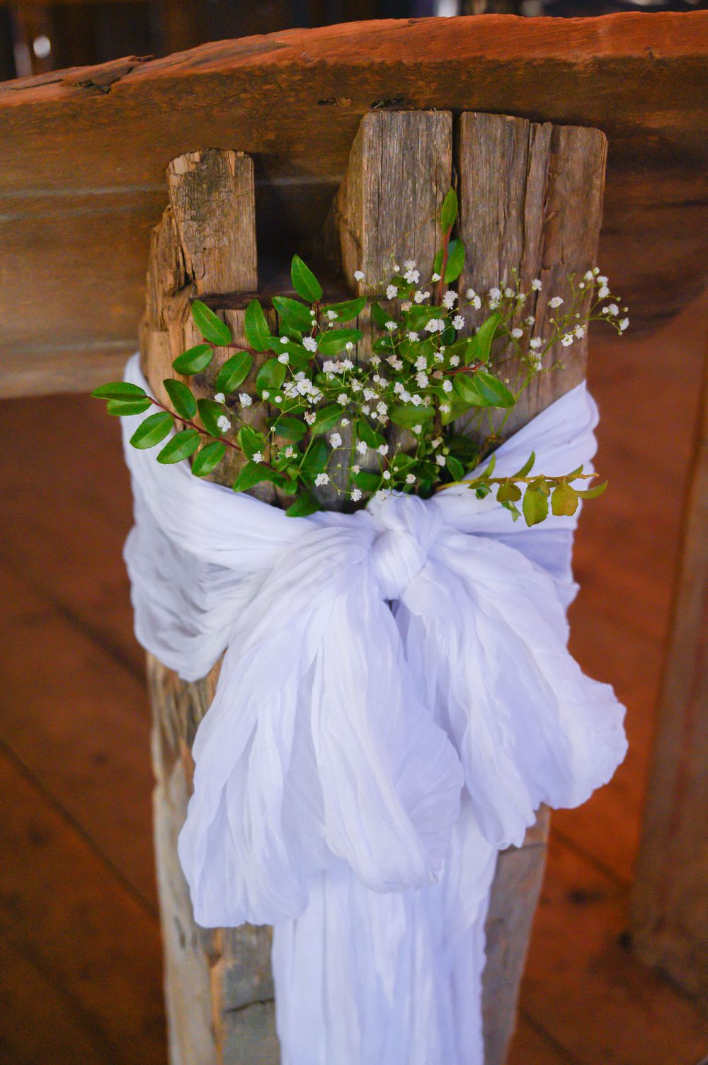 White satin bows and delicate floral arrangements adorn rustic wooden posts in a wedding ceremony setup.