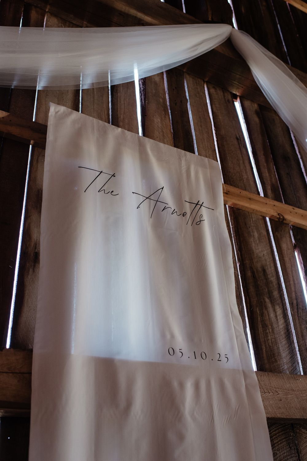 A white fabric banner with elegant script text and a date hangs from rustic wooden beams draped with sheer white fabric.