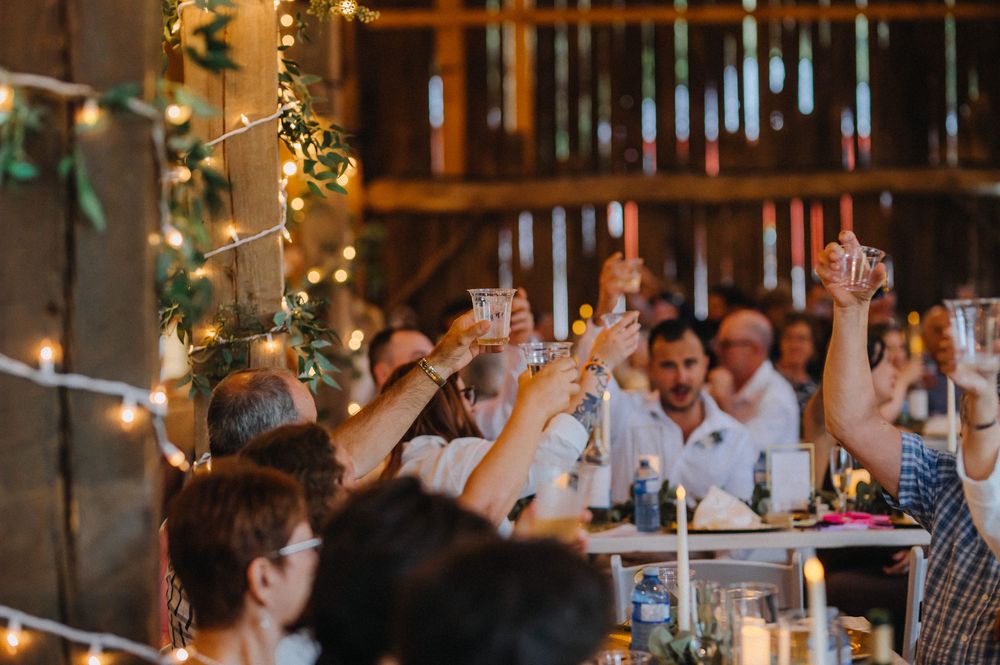People raise their glasses in a toast during a festive celebration with twinkling lights in a rustic indoor venue.