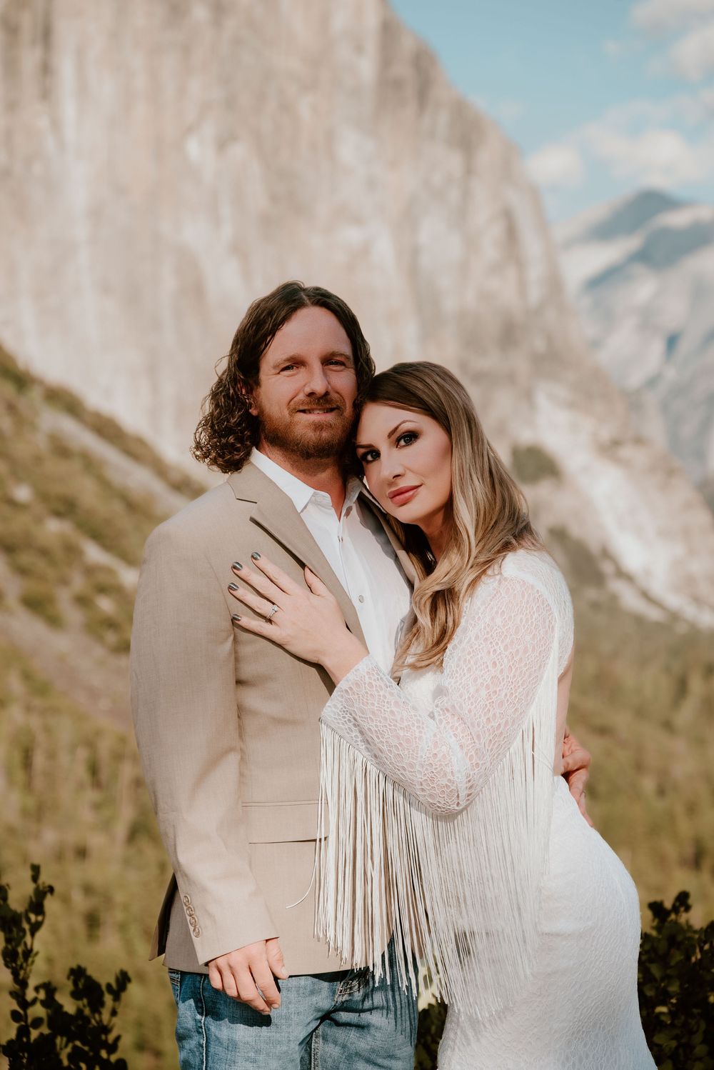 A couple in romantic embrace wearing white and beige against mountainous backdrop in natural lighting.