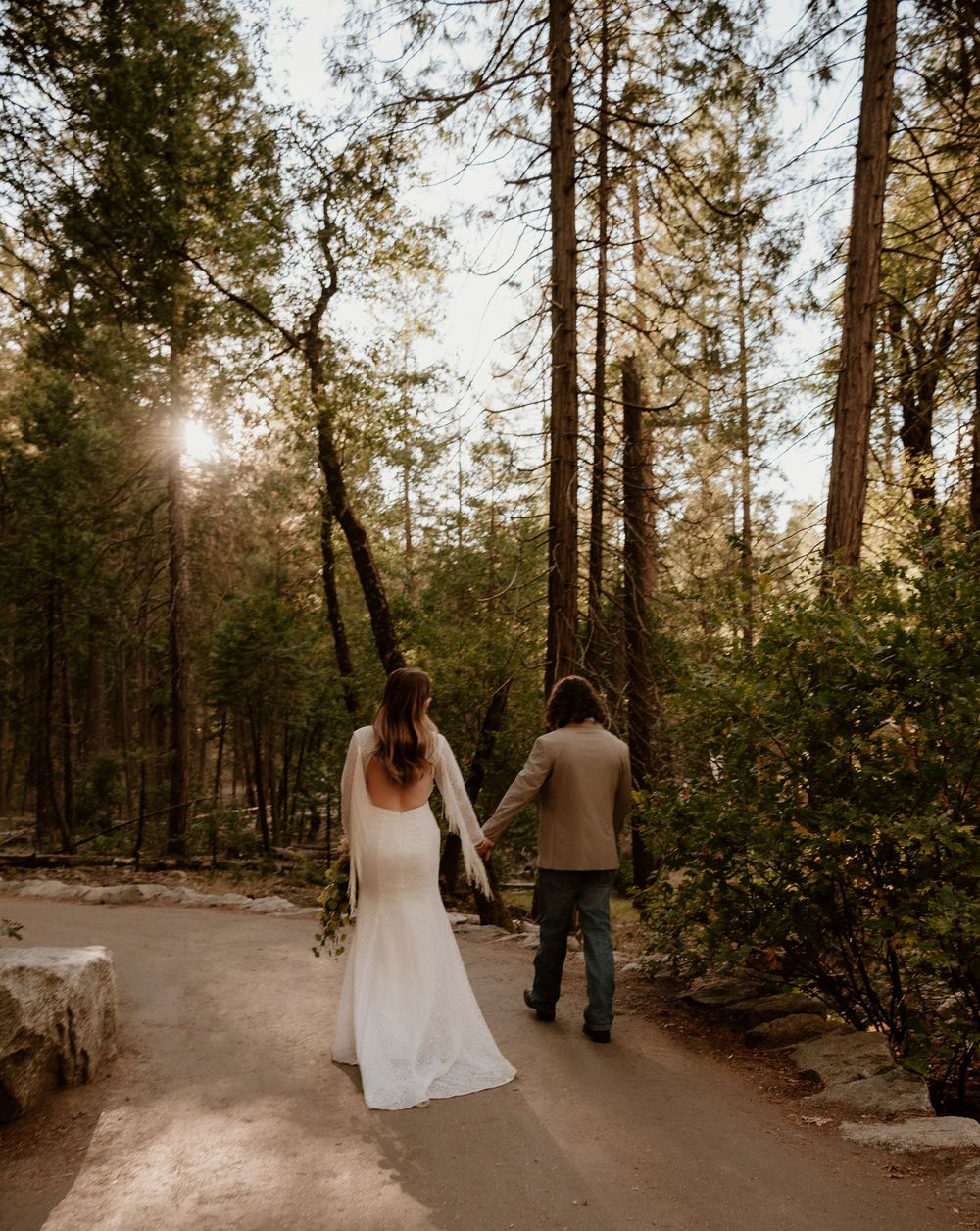 A couple walks hand in hand along a sunlit forest path in Yosemite National Park during golden hour.