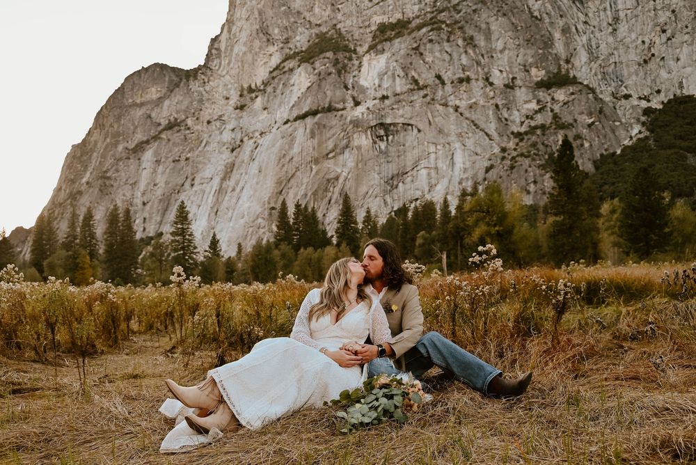 A romantic couple sits in a meadow with a stunning granite cliff face of Yosemite Valley in the background.