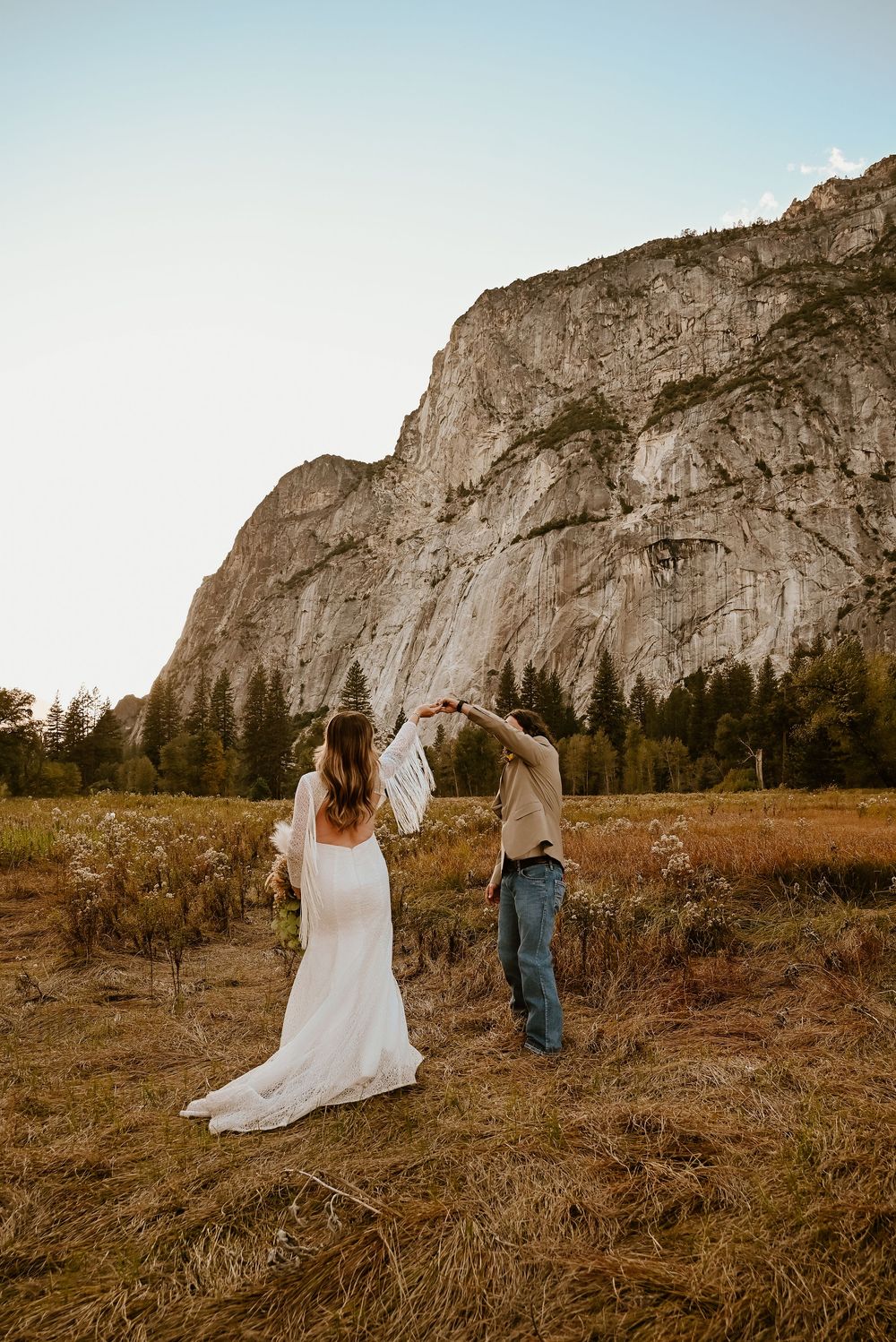 A couple walks through a meadow at sunset with towering granite cliffs of Yosemite Valley in the background.