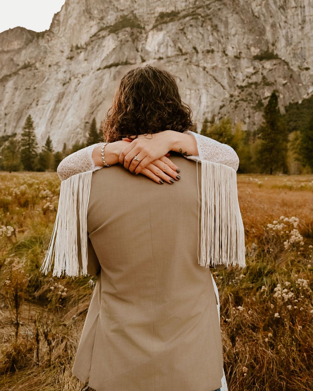 Person wearing fringe jacket stands in meadow with dramatic granite cliffs of Yosemite Valley in background.
