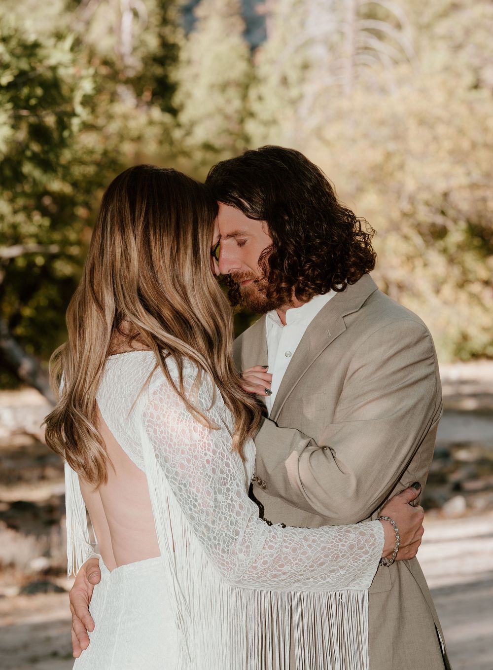 A romantic outdoor photoshoot shows a couple in a white dress and beige suit embracing in a sunlit forest setting.