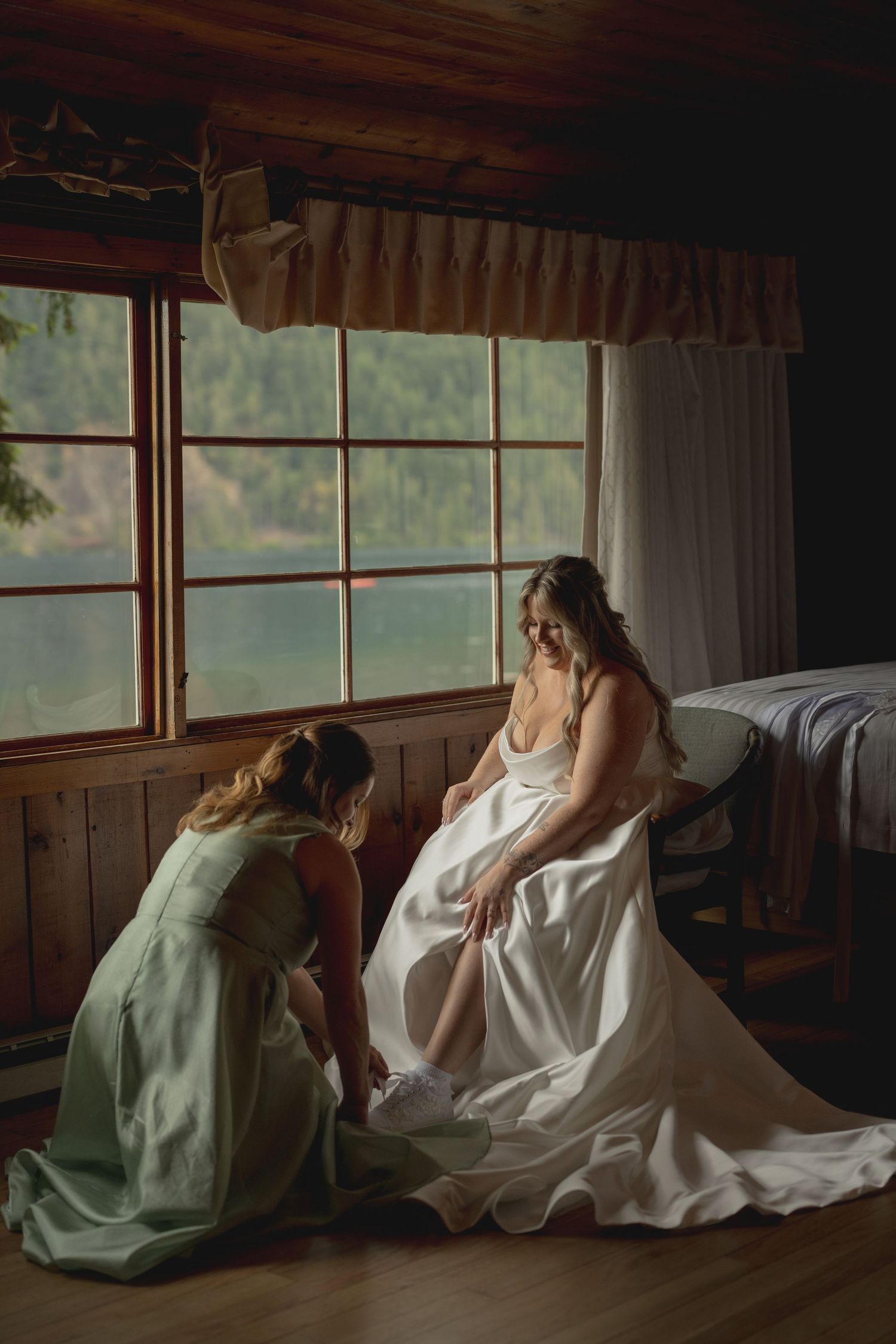 Bridesmaid helps bride with shoes in a rustic cabin room with large windows on wedding morning.