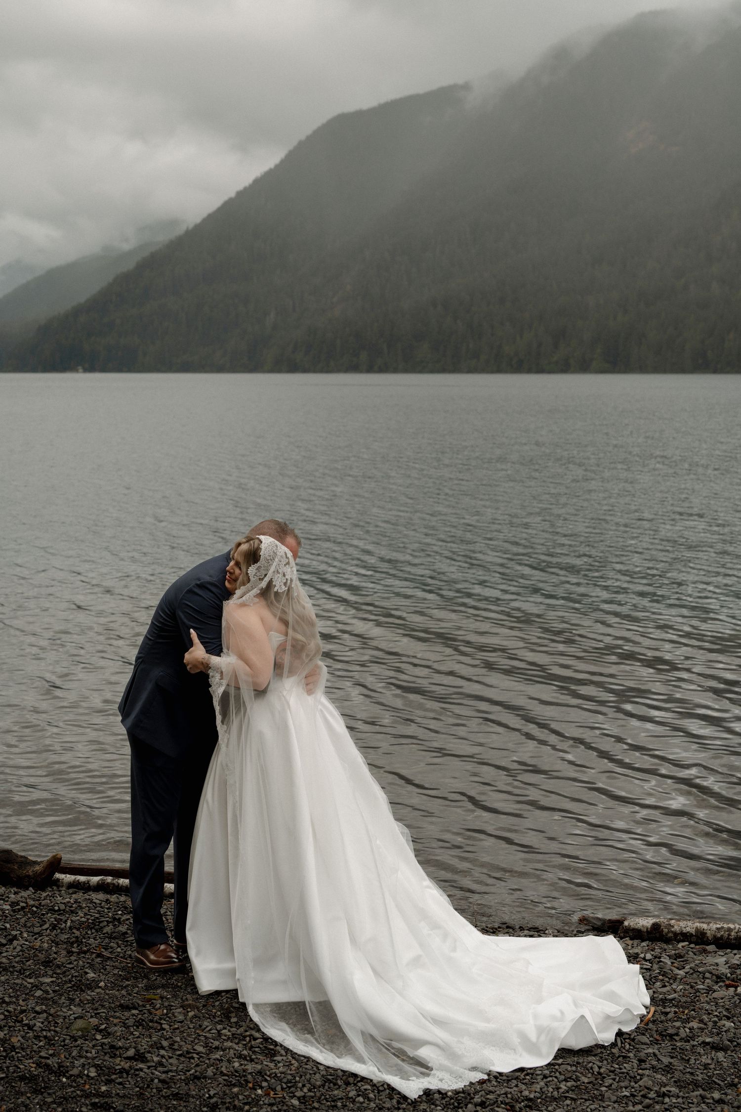 Bride and groom share a kiss by a misty mountain lake, her white gown trailing on the rocky shore.
