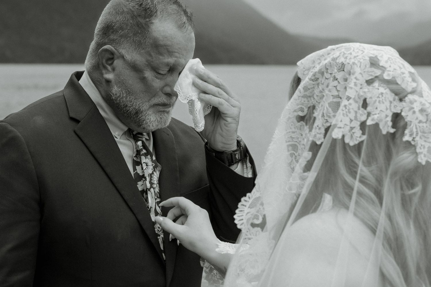 Emotional black and white wedding photo of father wiping tears while touching bride's lace veil.