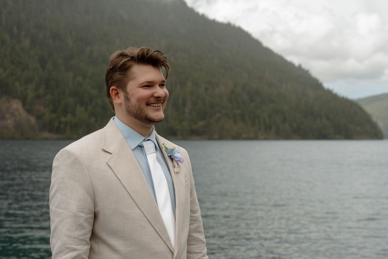 Smiling groom in tan suit with floral boutonniere stands before a scenic mountain lake backdrop.