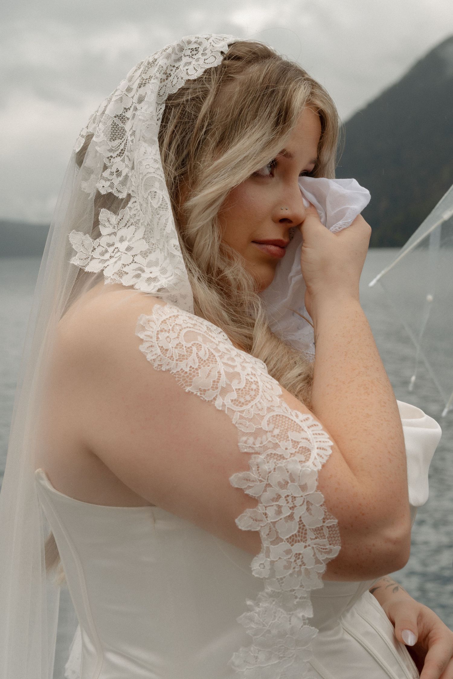 Emotional bride in lace veil wipes tears on a boat with mountain lake backdrop on her wedding day.