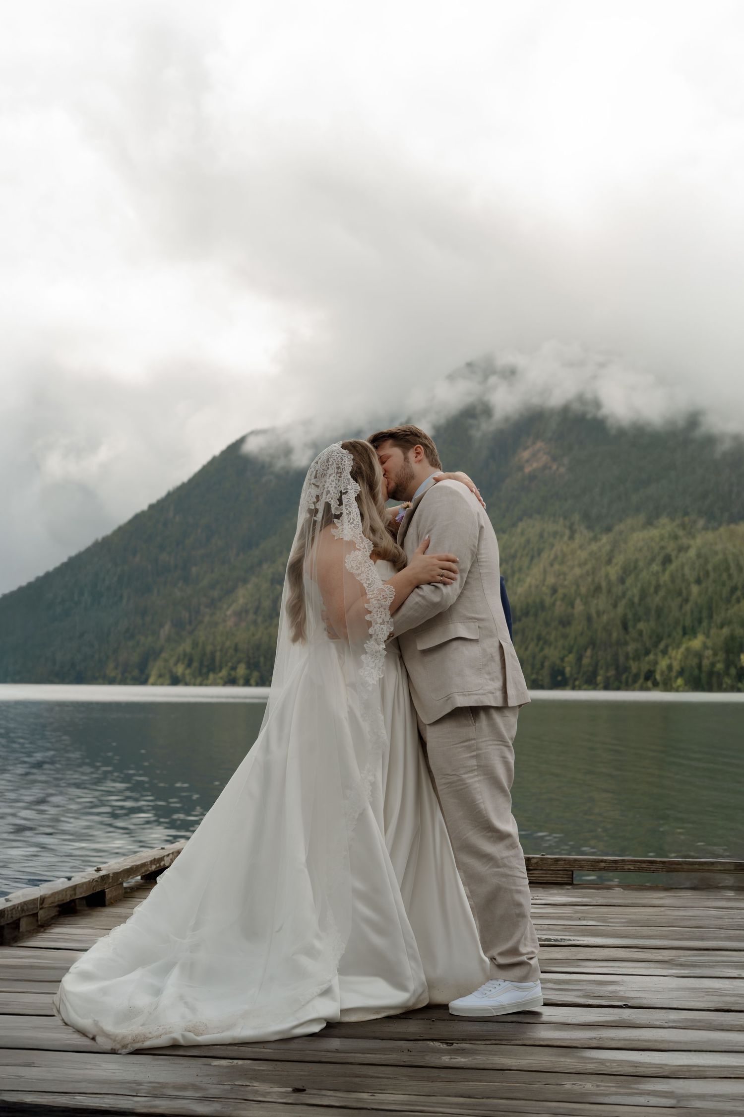 Bride and groom kissing on wooden dock with misty mountain lake backdrop on their wedding day.
