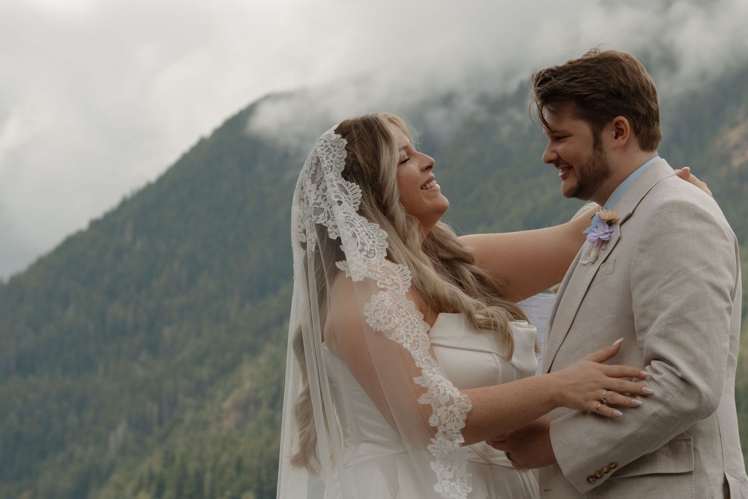 Bride and groom embrace outdoors with misty mountain backdrop on their wedding day.