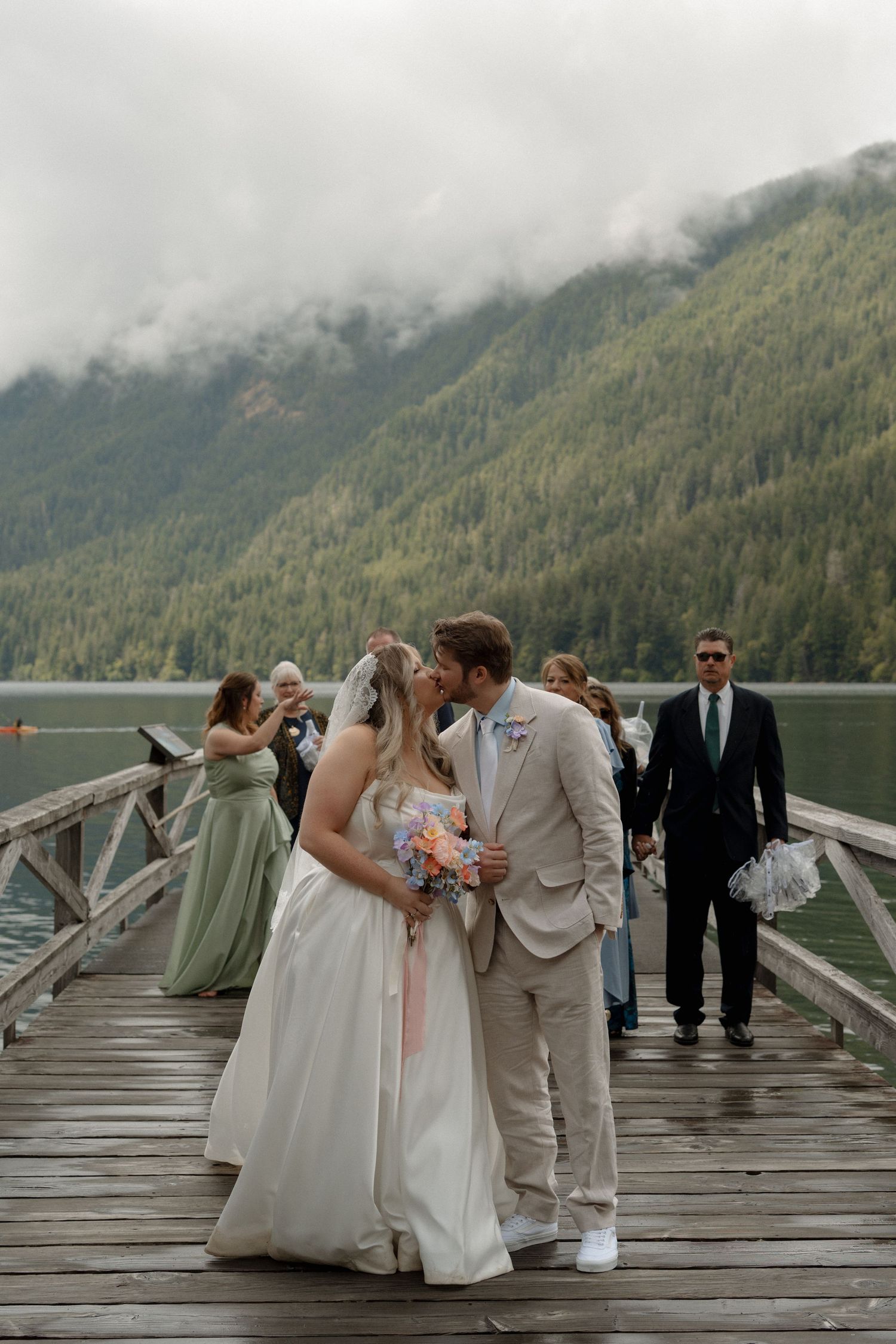 Bride and groom kiss on a wooden dock with misty mountain lake backdrop and wedding party.