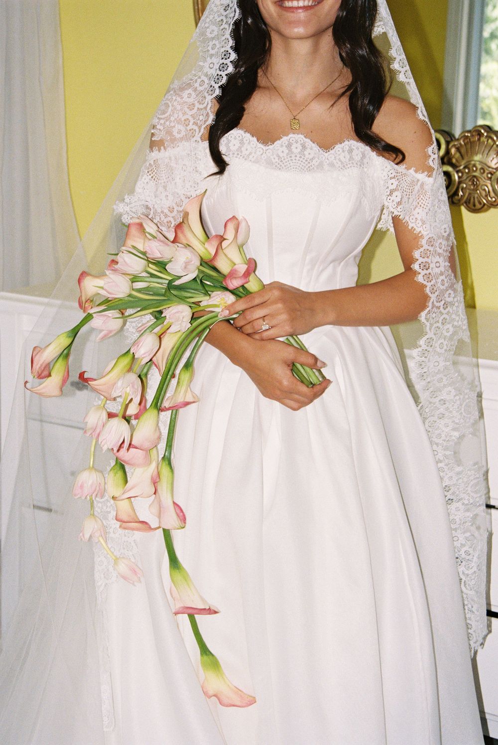 Bride in white off-shoulder gown with lace veil holding cascading pink calla lily bouquet, smiling elegantly.