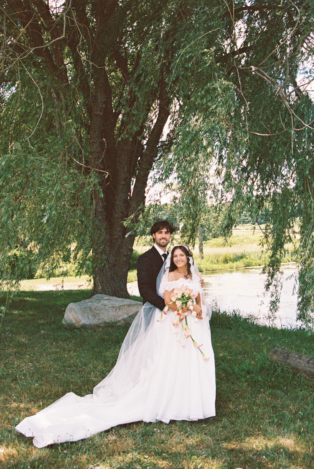 Bride and groom pose together under a weeping willow tree by a lake, bride holding a bouquet in a white gown.