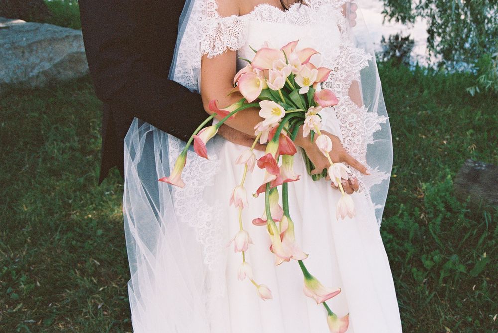 Bride in white lace wedding gown holding cascading pink calla lily bouquet standing with groom outdoors.