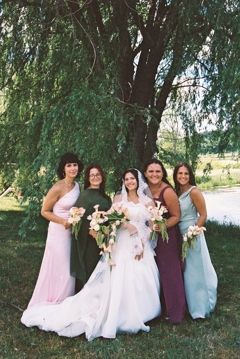 Bride in white gown poses with four bridesmaids in colorful dresses holding bouquets under a weeping willow tree.