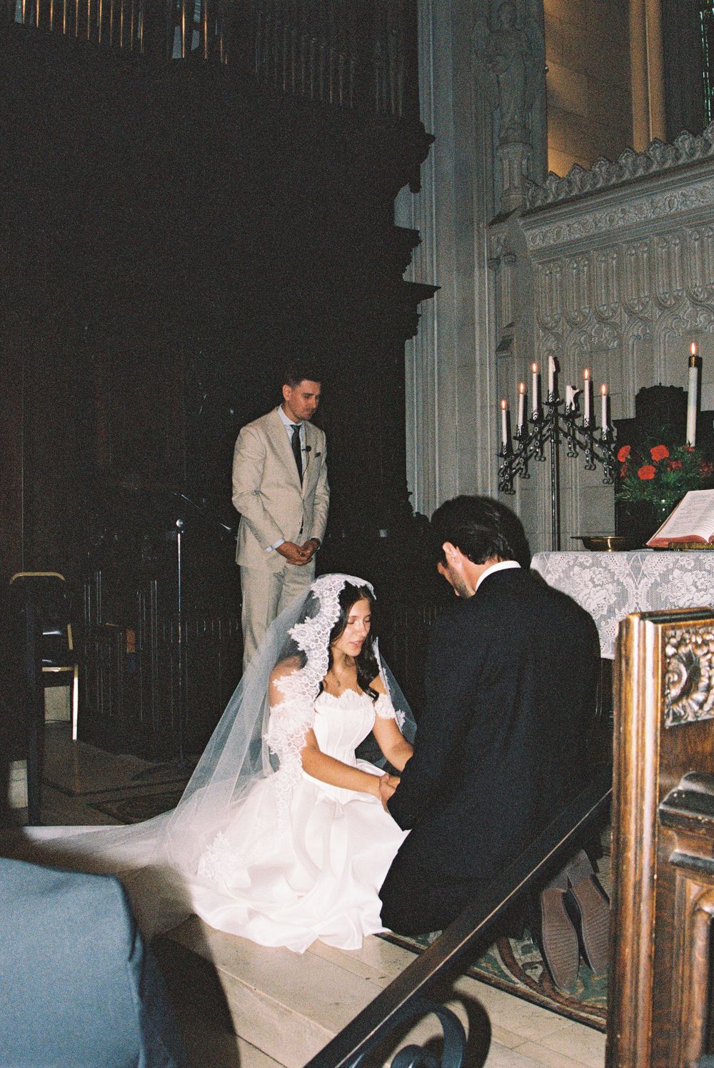 Bride in white gown and veil kneels at church altar during wedding ceremony with groom and officiant.