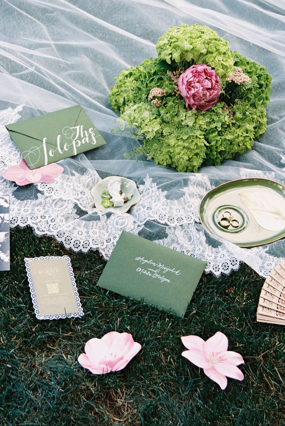 Elegant wedding stationery flat lay with green envelopes, rings, pink flowers, and lace veil on grass.