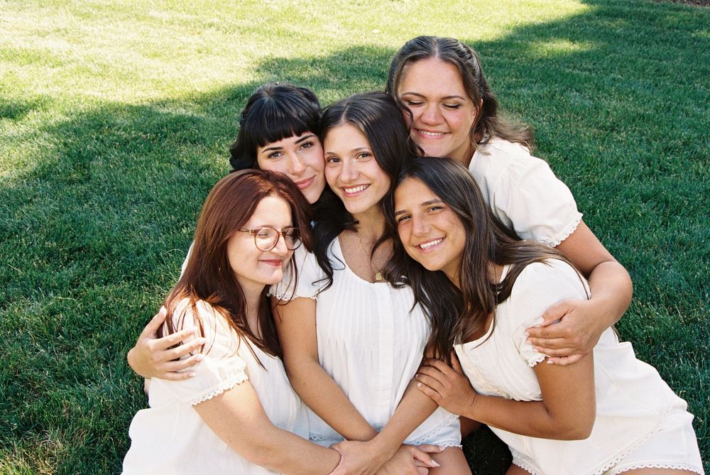 Five young women in white dresses smiling and posing together on green grass outdoors.
