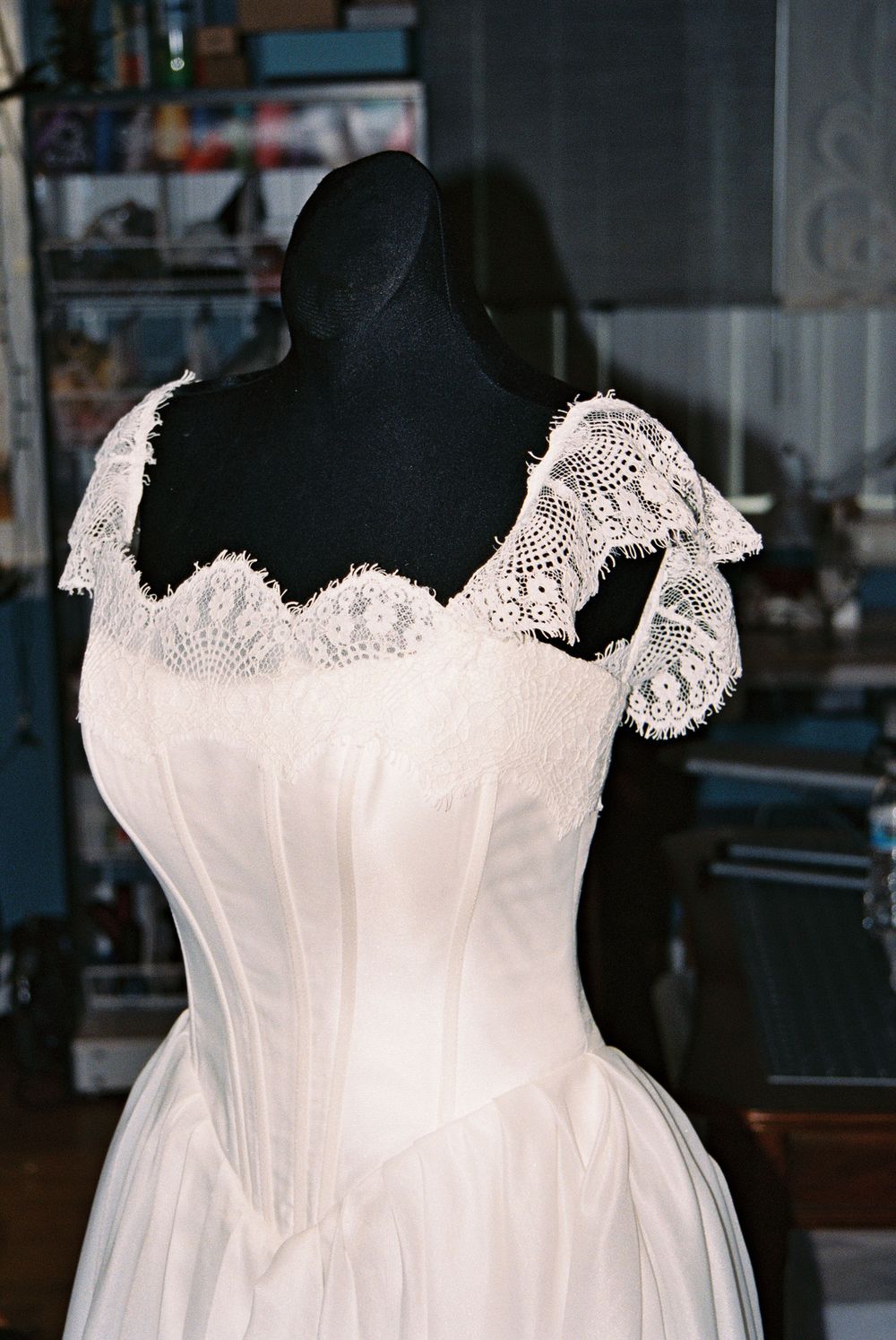 White lace wedding dress displayed on a black mannequin in a bridal boutique, featuring corset bodice and cap sleeves.
