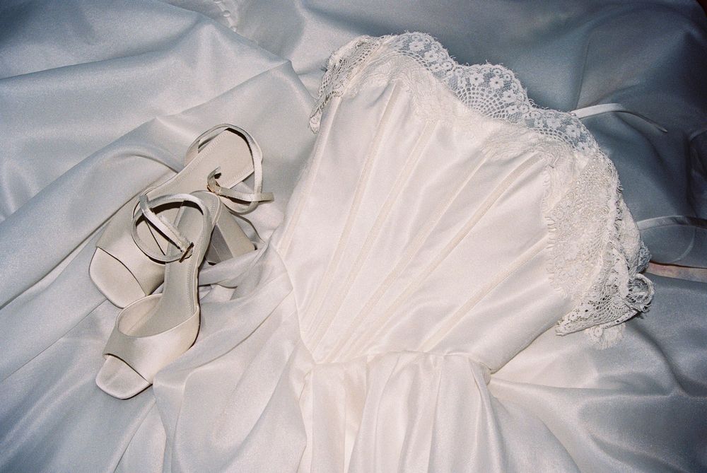 White wedding dress with lace trim and white strappy heels laid out on satin fabric.