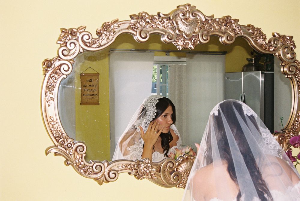 Bride in wedding veil adjusts her jewelry while gazing into an ornate gold baroque mirror.