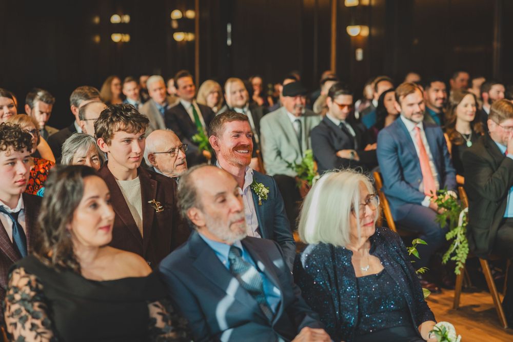 Wedding guests gathered in formal attire watch attentively during an indoor ceremony.