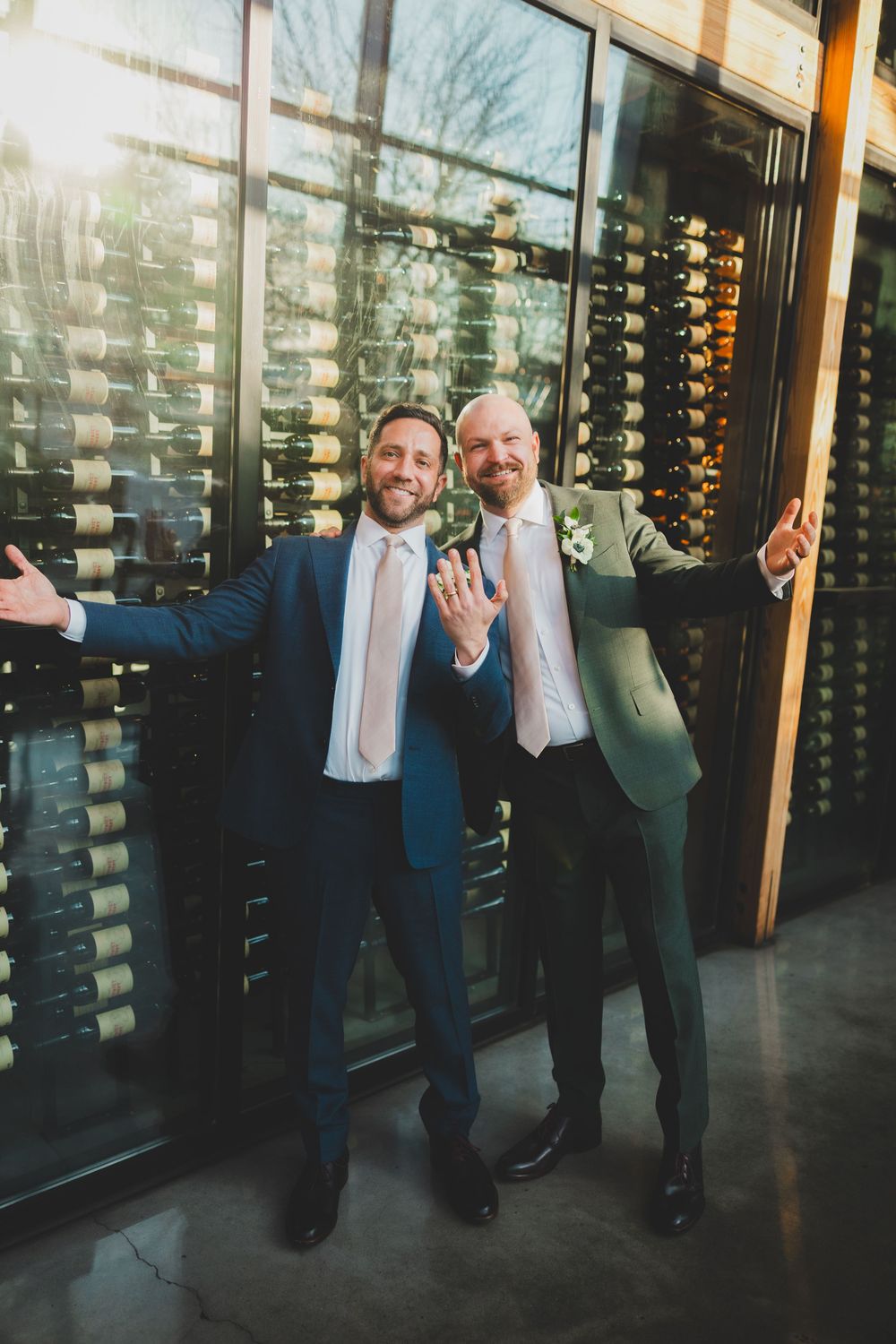 Two grooms pose playfully in front of a modern wine storage wall display in DC