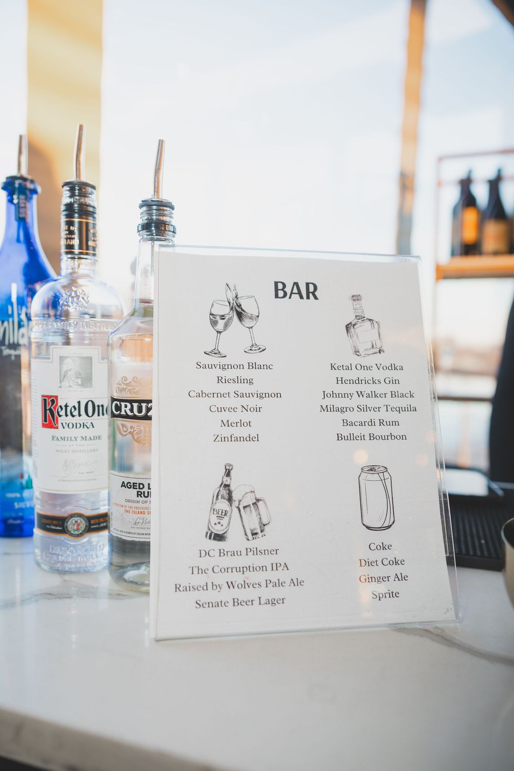 A row of liquor bottles and drink menu cards displayed on a white bar counter at an event or restaurant.