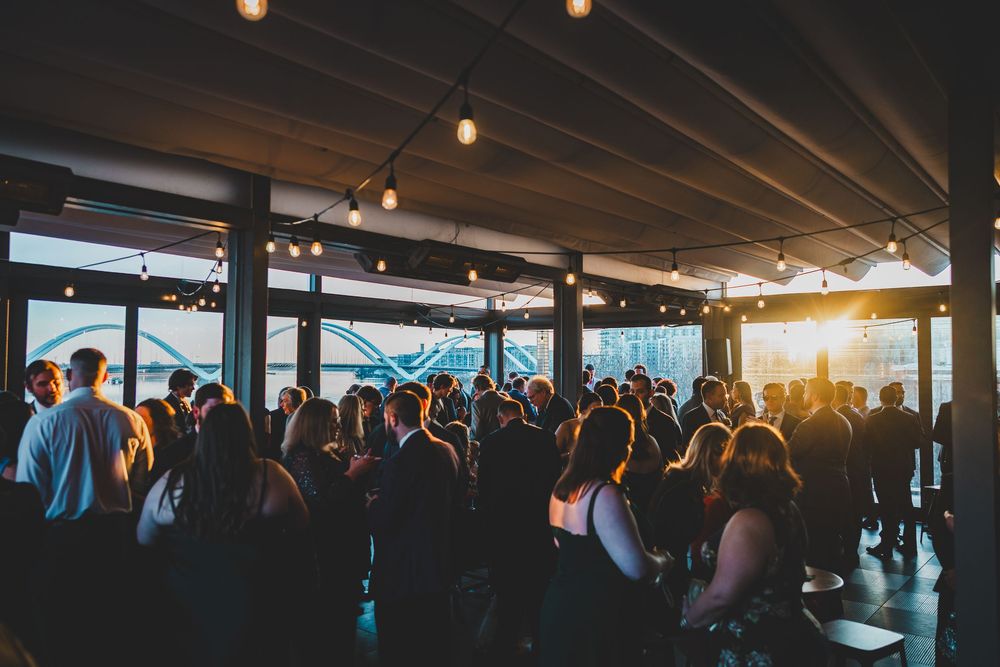 A crowded sunset event on a covered patio with string lights and ocean views in the background.