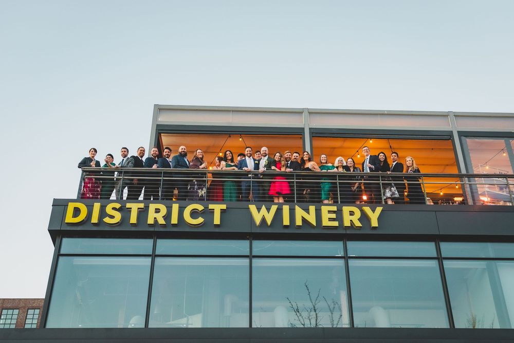 A crowd of people gathered on the balcony of District Winery, a modern glass building with illuminated orange signage.