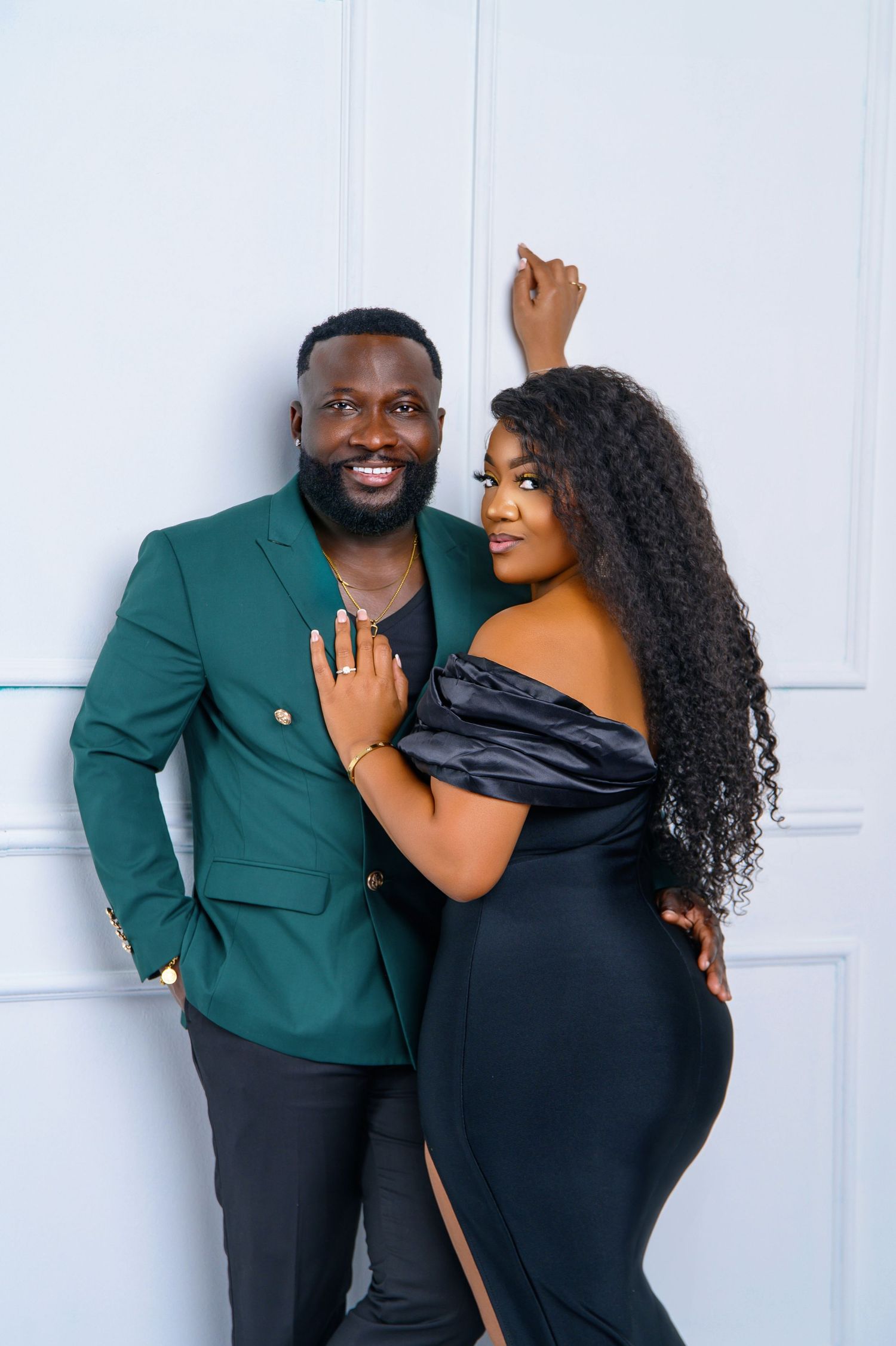A couple poses together against a white wall, wearing green and black formal attire.