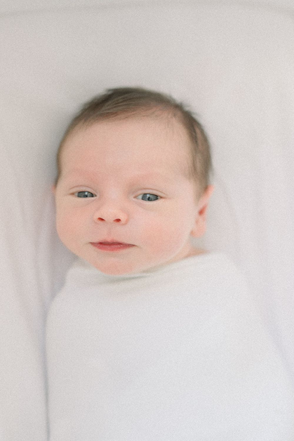 A sweet baby with rosy cheeks rests peacefully on soft white bedding, gazing upward with bright eyes at an in-home lifestyle session in DC