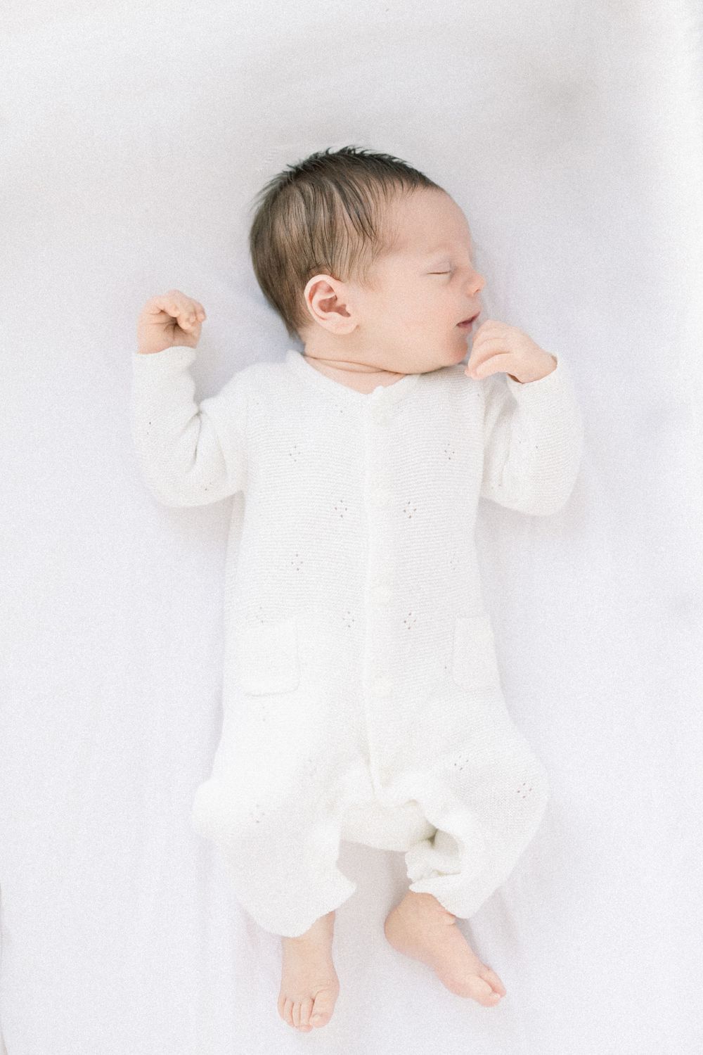 A peaceful sleeping baby wearing white clothing lies on soft white bedding at his lifestyle newborn session in Washington DC