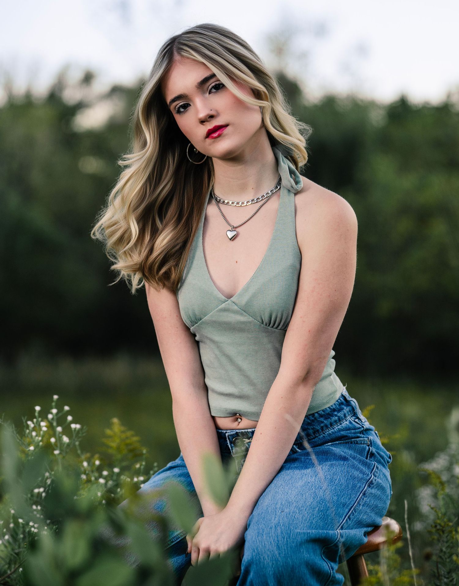 A person in a sage green tank top and blue jeans poses outdoors in a field with wildflowers at golden hour.